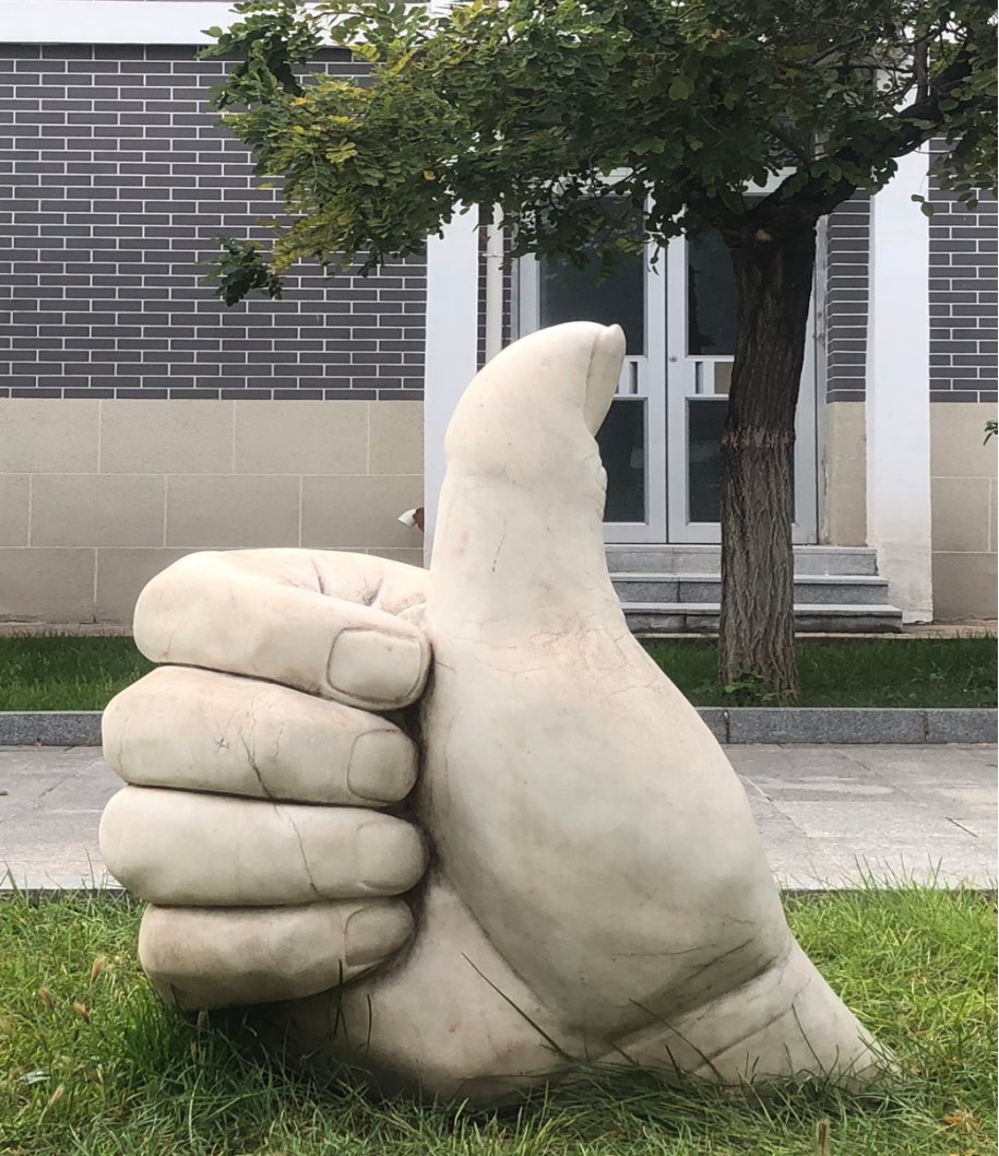 校园绿化带中的真实大拇指雕塑,后方是树木与校园建筑。