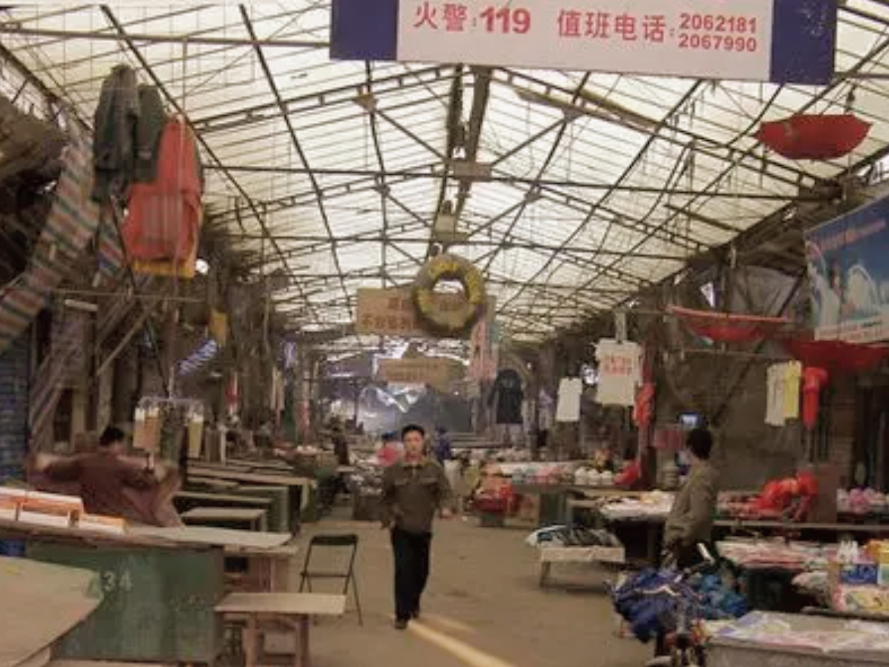 相似环境参考,非原始现场照片:有顶棚的市场通道,两侧摊位摆放着货物,有人在通道中行走。